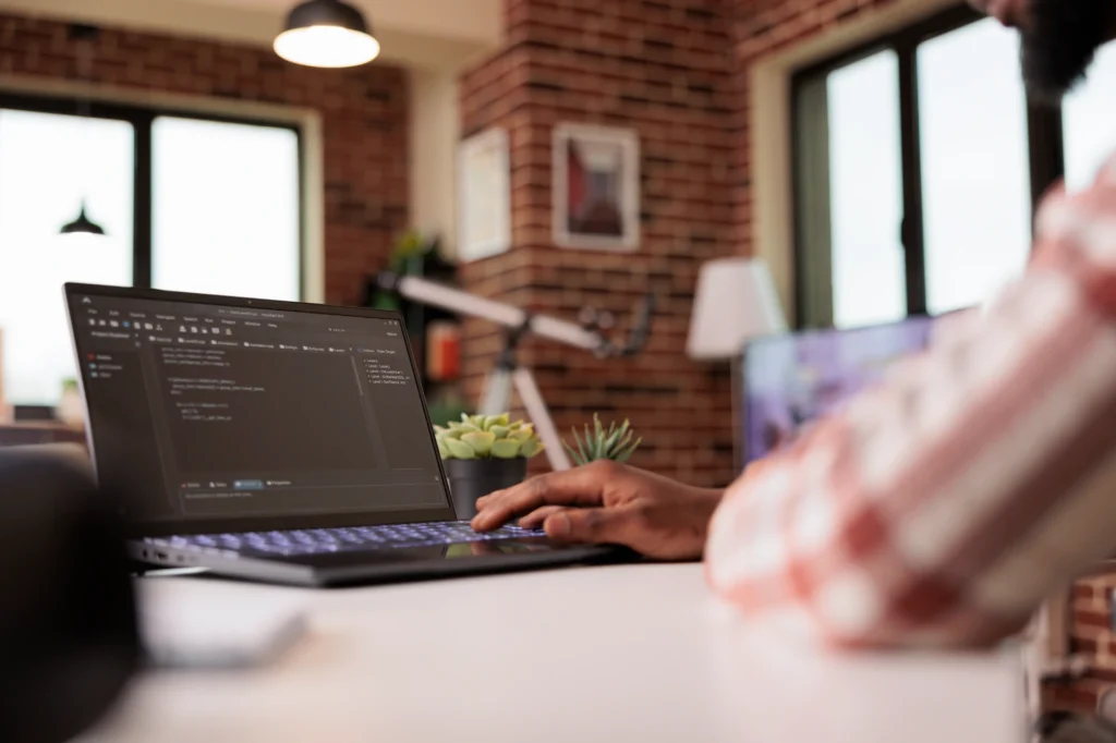 Developer typing on a laptop in a brick-walled home office with blurred background.