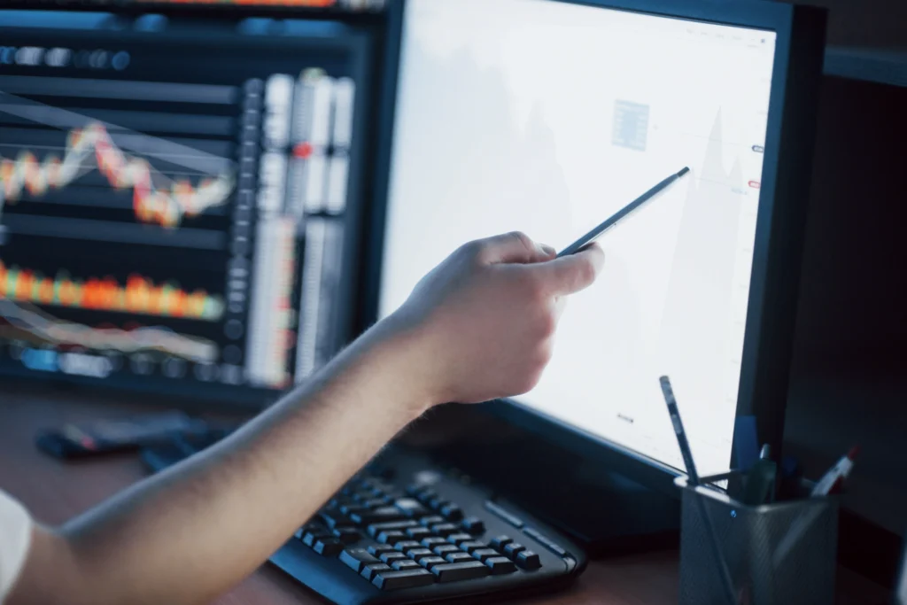 Analyst pointing a stylus at a line chart on a monitor beside trading graphs.