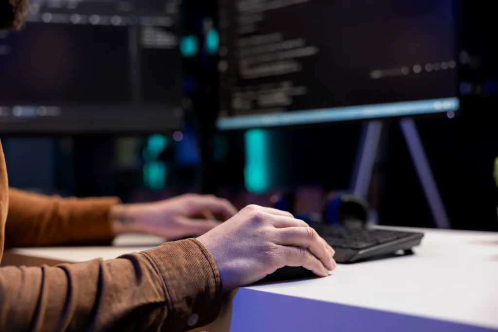 Developer at a desktop keyboard with code on twin monitors in a dim workspace.