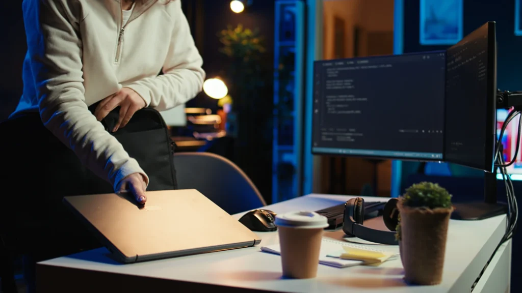 Person packing a closed laptop into a bag on a desk with code visible on dual monitors.