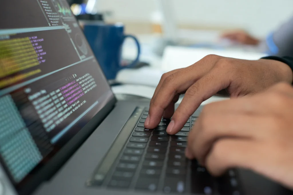Close-up of hands typing on a laptop terminal full of colorful code lines.